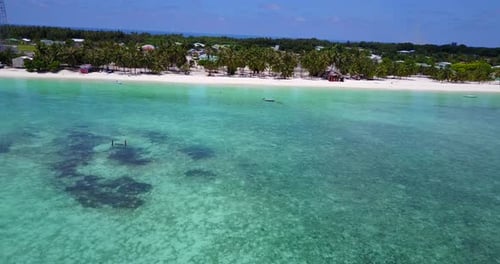 Low aerial along Maldives island coast with shallow clear water. Houses, resorts and palm trees by w