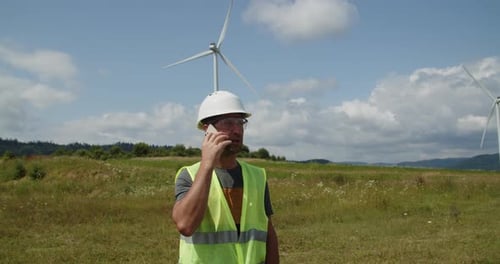 Engineer Caucasian Man in Hardhat and Goggles Talking on a Cell Phone to Discuss Wind Turbine