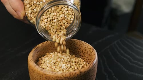 Pouring Raw Whole Grain Green Buckwheat Seeds From Glass Jar Into a Wooden Cup