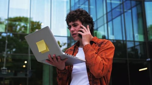 Man Talking on Cell Phone Pacing Back and Forth Outdoors Near Office with Laptop in His Hands