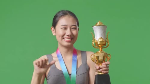 Asiatische Frau zeigt der Kamera eine Goldtrophäe, lächelt und zeigt sich selbst auf einen Greenscreen-Hintergrund
