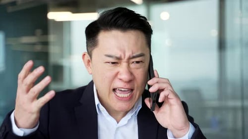 Angry asian businessman arguing talking on mobile phone at workplace in office. Excited nervous
