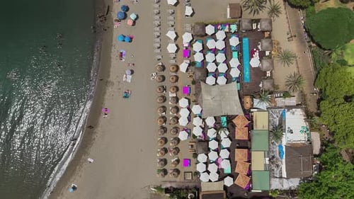 Top-down drone view of beach resort with umbrellas, sunbeds and resting people by the shoreline in