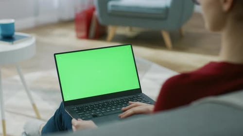 Young Woman at Home Works on a Laptop Computer with Green Mock-up Screen. She's Sitting On a Couch