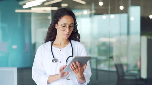 Confident female doctor in a white coat is using digital tablet in modern hospital clinic.