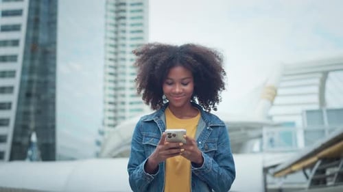 Happy young African American woman walking down the city street smiling using mobile phone chatting