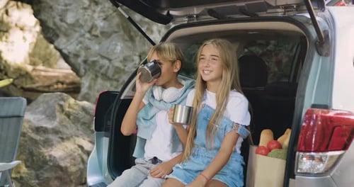 Cheerful brother and sister sitting in car trunk and drinking tea and speaking during spring trip