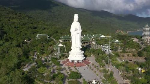 Close aerial circling around tall Lady Buddha statue and temples with huge mountains and ocean in Da