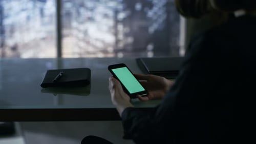 Over the Shoulder View of Businesswoman Sitting at Her Desk and Using Smartphone with Green Screen.