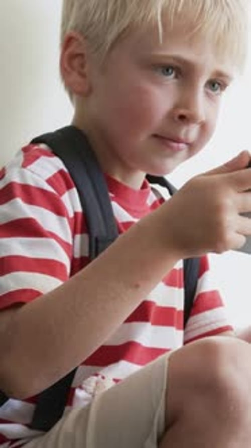 Vidéo verticale : Enfant assis en t-shirt rayé jouant sur une tablette près d'une fenêtre lumineuse montrant un sac à dos