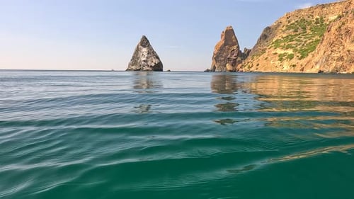 Sea Water Surface Camera Flies Over the Calm Azure Sea with Volcanic Rocky Shores on Background