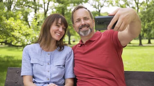 A Middleaged Caucasian Couple Takes Selfies with a Smartphone and Smiles on a Bench in a Park