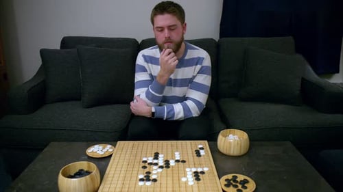 Beaded man strokes beard while thinking of strategic move in Go board game on bamboo board on stone