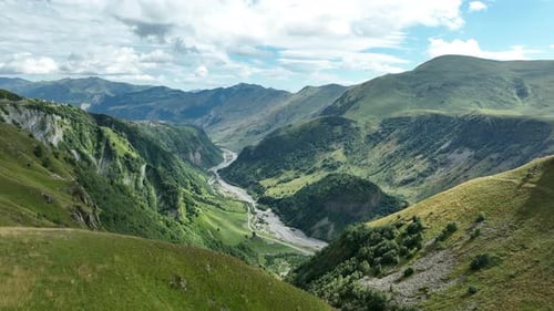 Aerial view of a deep mountain gorge with a winding riverbed, lush green slopes, and rugged peaks