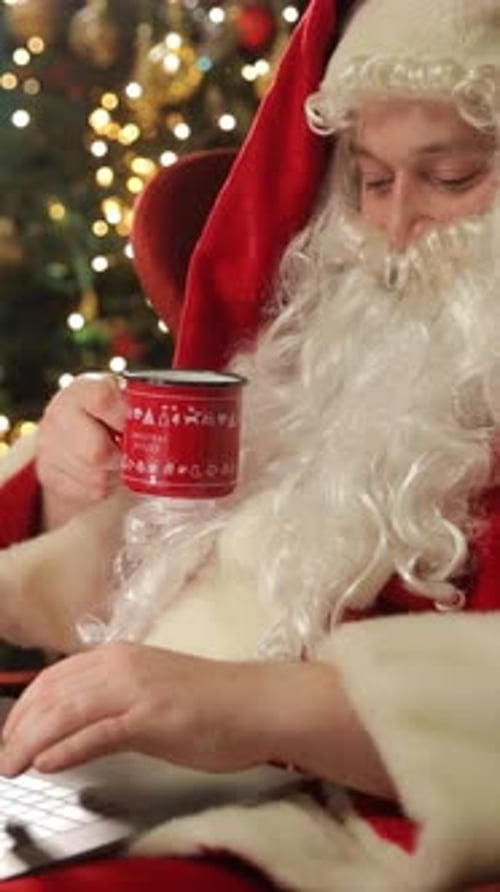 Santa Claus Drinks From a Mug While Working on a Laptop in a Festive Room Vertical Video