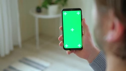 Young Man Sitting at Home Holding Smartphone Green Mockup Screen in Hand Male Person Using Chroma