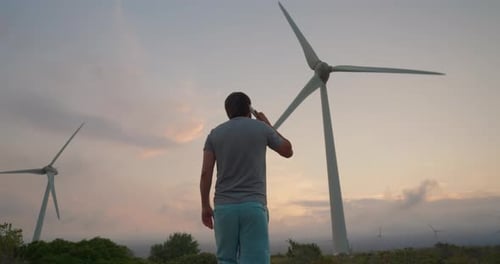 Man Standing Against Large Windmill on Sunny Day Talking on the Phone