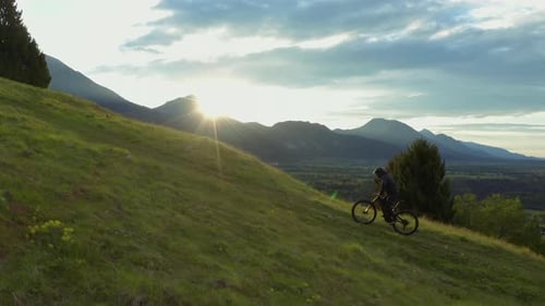 Young athletic man on MTB e-bike pedalling up the hill with green grass at sunset/sunrise.