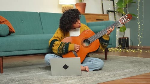 Woman Plays Guitar and Watches Laptop Indoors