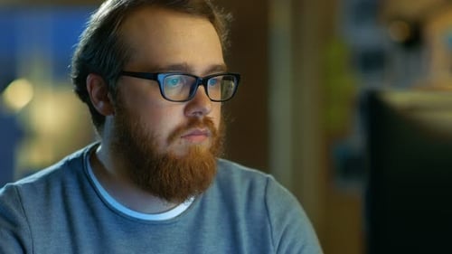 Young Bearded Creative Man With Glasses Thinks on a Problem While Working on His Personal Computer.