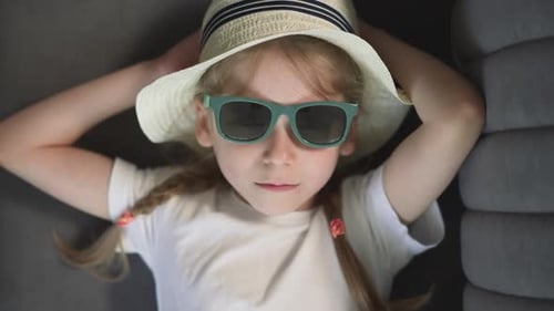 Sad Little Girl in Sunglasses with Green Rim Lies on Sofa