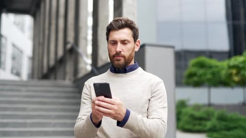 A serious businessman is using a phone while standing on the street near an office building.