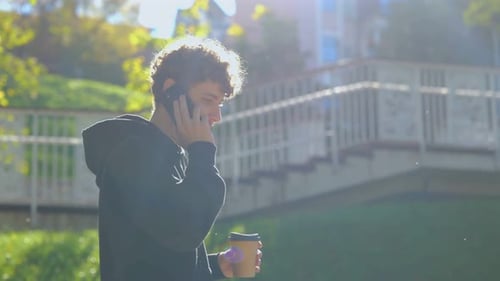 Happy Young Man Talking on the Mobile Phone While Walking in the Park and Holding Cup of Coffee