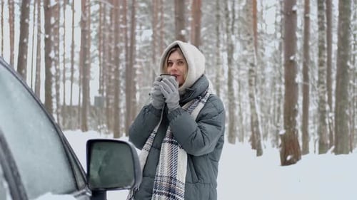 smiling woman drinks hot tea in the winter forest near the car