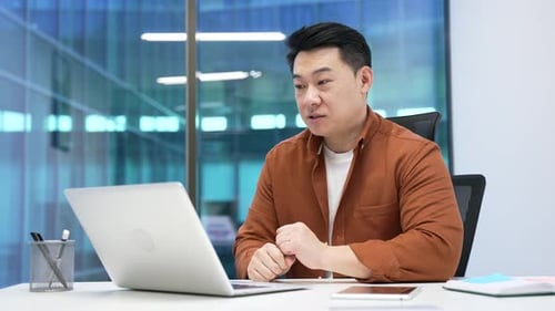 Confident asian businessman talking on a video call using laptop sitting at workplace in modern