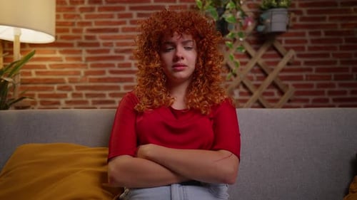 Young woman with curly red hair sitting on a couch, arms crossed, looking sad.