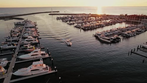 A serene sunset view over a bustling marina with boats docked and a lone vessel cruising