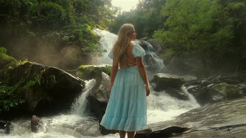 Frau in fließendem blauem Kleid steht am ruhigen Wasserfall in üppigem Grün
