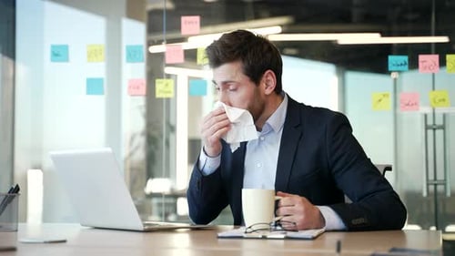 Sick businessman suffers from a cold while sitting at a desk at a workplace in a business office.
