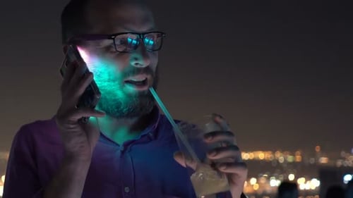 Young man enjoys cocktail and cellphone chat on bar terrace at night