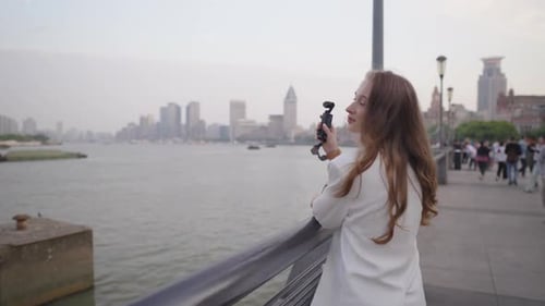 Confident Female Journalist in White Blazer Holding Microphone Standing on Riverside Promenade with