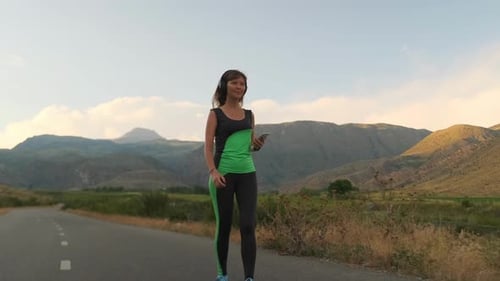 A Young Woman Walks a Deserted Road Switching Music Maintaining Fitness