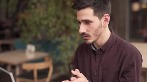 Young man is sitting in a cafe in front of the laptop and talking with somebody in a video chat. Man