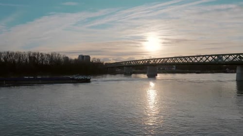 View of wide river with bridge spanning over it and small cargo vessel passes by at sunset
