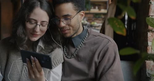 Happy Biracial Couple Looking at Smartphone Screen Talking Sitting in Cafe Together