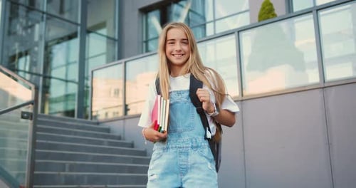 Cheerful smiling cute caucasian schoolgirl with backpack standing on stairs near school posing
