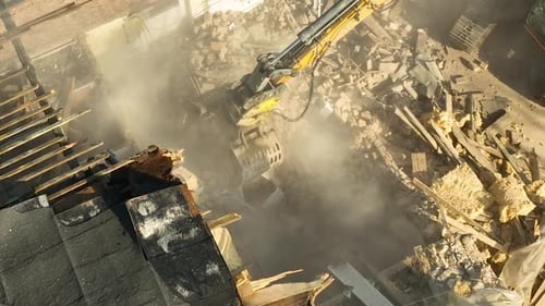 Aerial view of a construction site where an excavator's arm is demolishing a building, creating dust