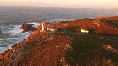 Rough sea waves hitting rocky coast with lighthouse complex on top, aerial orbit view