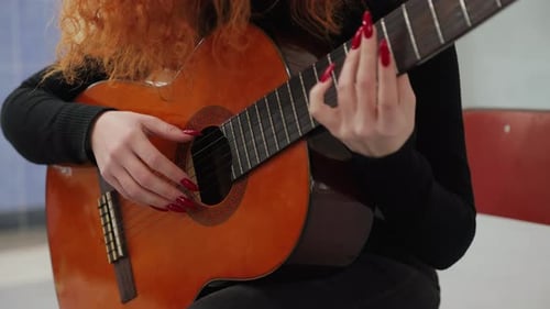 Close-up video of a woman playing chords on an acoustic guitar while sitting on a stool in a room.
