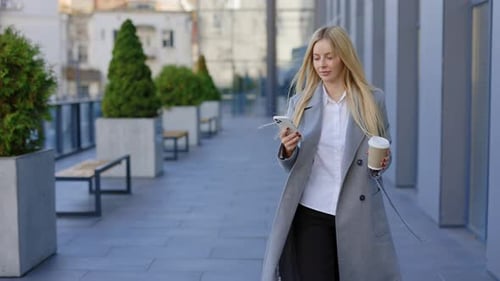 BlondeHaired Businesswoman Leisurely Walking on the Street Holding a Mobile Phone and a ToGo Coffee