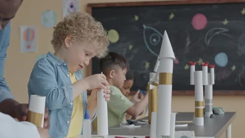 Boy Making Paper Rocket with Help of Teacher at School