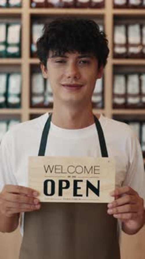 Portrait of Cheerful Waiter in Uniform Showing Sign Welcome Open in Cafe