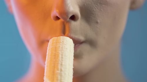 Close Up Of Woman's Mouth Eating Banana Fruit, Studio Shot With Colorful Lighting