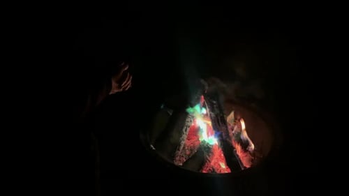 Camper on dark black night warms hands by colorful camp fire flames