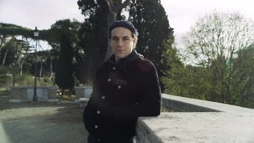 Portrait Of A Content Italian Man Leaning Against A Stone Ledge In A Park With Trees In The Backg...