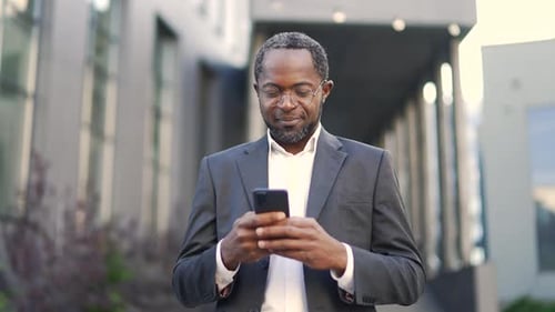 Mature African American businessman standing outdoors in front of office building using smartphone.